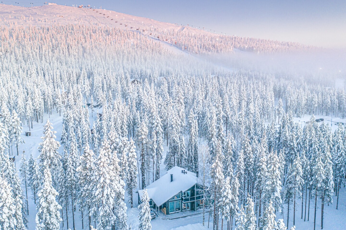 Aerial view of a chalet surrounded by snow-covered evergreen trees, with a ski slope in the distance and a pinkish glow over the mountain